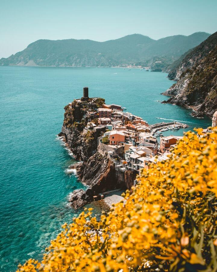 Auf dem Bild ist Cinque Terre im hintergrund eines Blumenfeldes zusehen, die Blumen sind gelb bis orange. Der Focus liegt auf der Stadt.