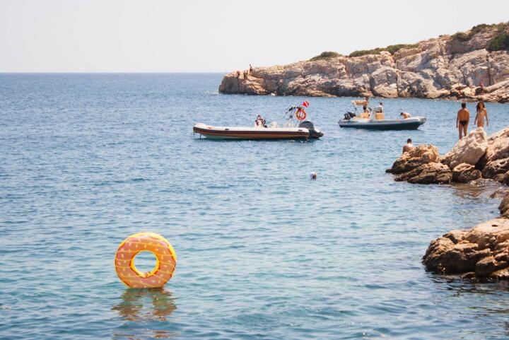 Ein schwimmring im Wasser, der Schwimmring symbolisiert einen Doughnut und steht aufrecht im Wasser. Im hintergrund sind 2 Schlauchbote und eine Steinige Küste zusehen.